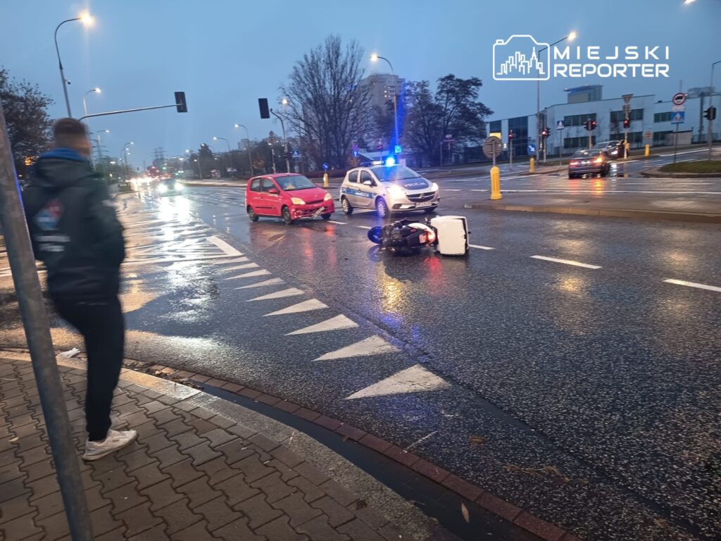 Zniszczony skuter leży na mokrej jezdni, obok uszkodzony czerwony samochód, a w tle widoczny radiowóz policyjny.