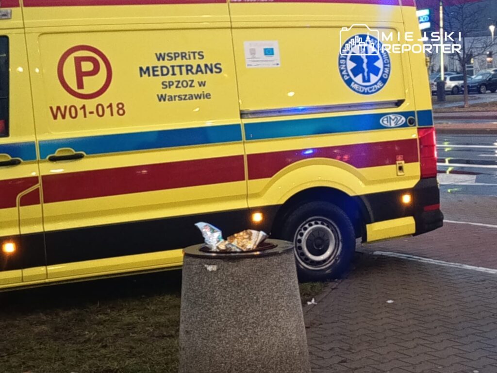 Karetka pogotowia medycznego zaparkowana przy chodniku obok kosza na śmieci, w tle nocne oświetlenie ulicy.