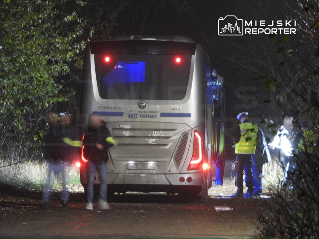 Grupa osób w odblaskowych kamizelkach sprawdza autobus na leśnej drodze w nocy.