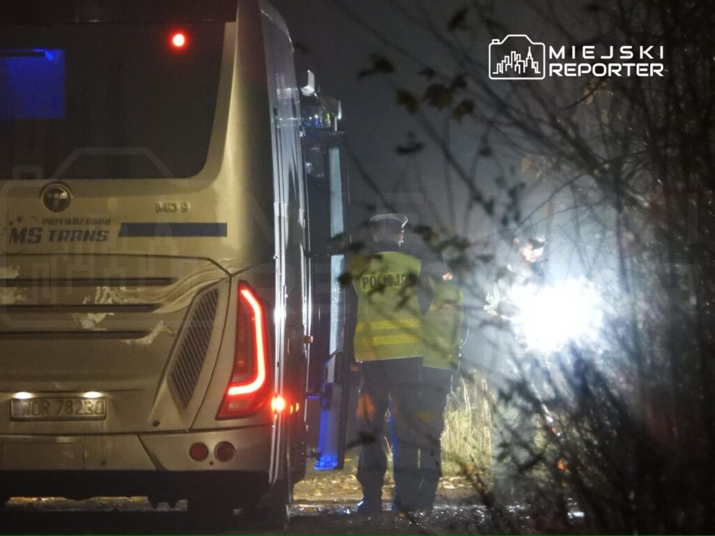 Policjanci w odblaskowych kamizelkach rozmawiają obok autobusu w ciemnym, leśnym otoczeniu.