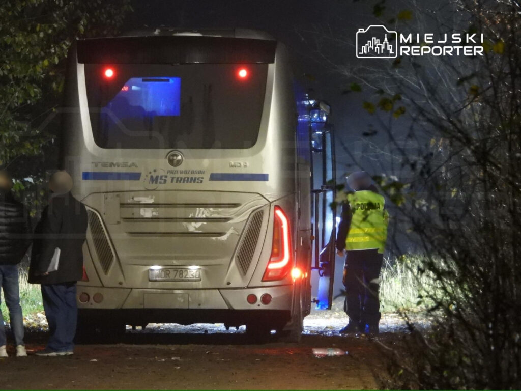 Grupa osób rozmawia obok autobusu z napisem "MS TRANS" na ciemnej, leśnej drodze, w pobliżu funkcjonariusza w kamizelce.