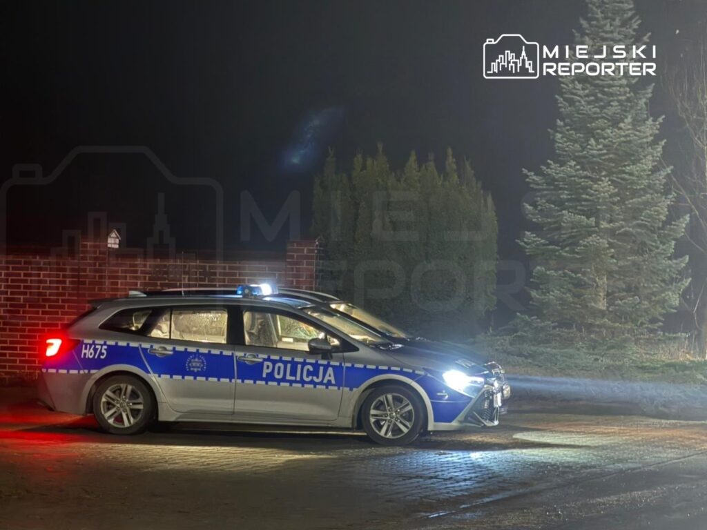 Policyjny radiowóz z niebieskimi światłami parkuje w zaciemnionym otoczeniu, obok drzew i ogrodzenia.