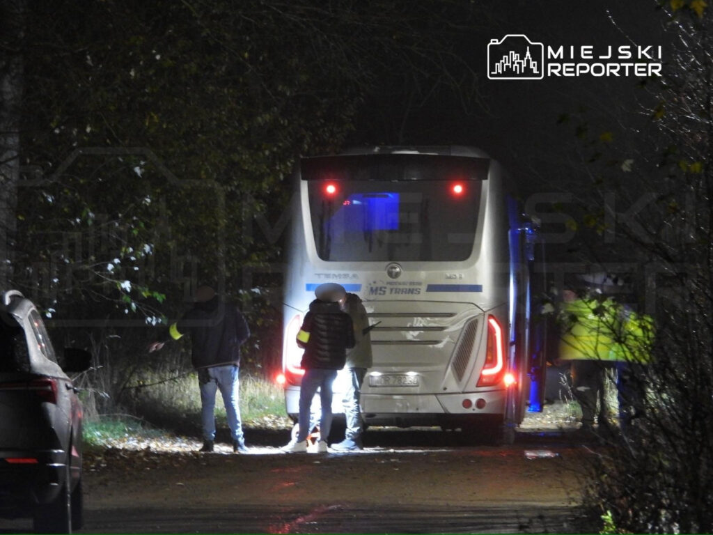 Grupa osób rozmawia obok zaparkowanego autobusu na oświetlonej drodze w nocy, otoczonej drzewami.