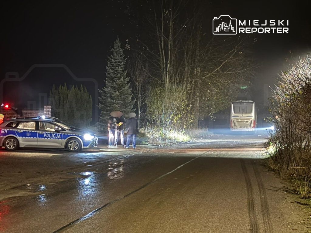 Policjant rozmawia z grupą osób na poboczu drogi w nocy, obok radiowozu z włączonymi światłami.