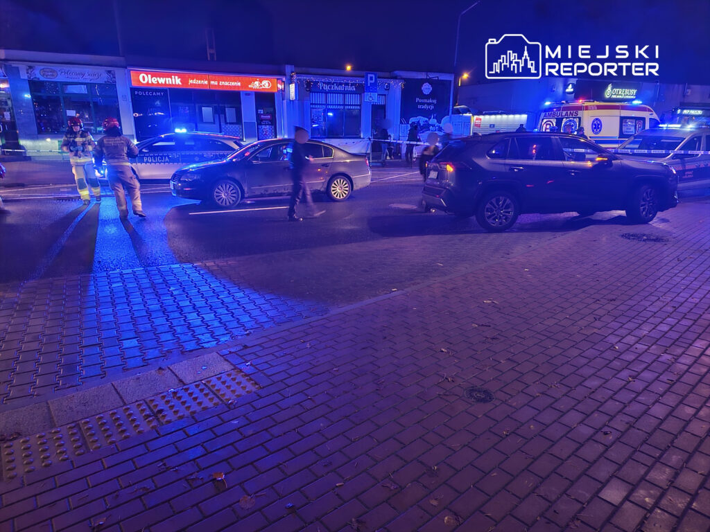 Na ulicy w nocy policjanci i strażacy interweniują przy wypadku, a w tle widać zaparkowane pojazdy oraz radiowóz.