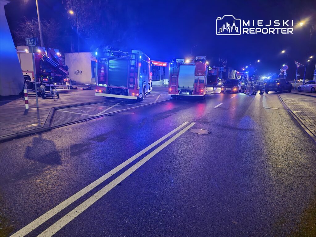 Na drodze zaparkowane są dwa wozy strażackie z włączonymi sygnalizacjami świetlnymi, a obok pracują strażacy.