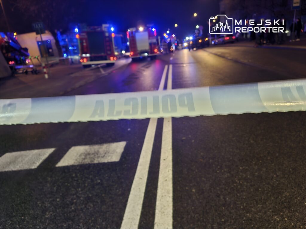 Policyjna taśma zabezpieczająca na jezdni, w tle widać pojazdy straży pożarnej i policyjne światła.