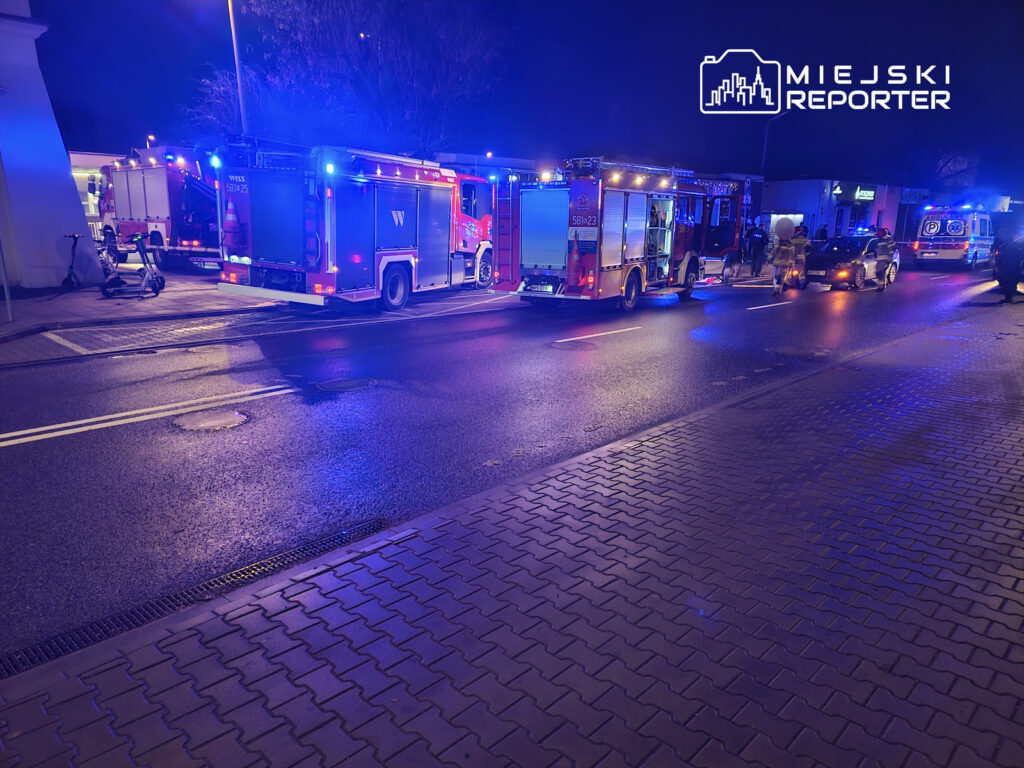 W nocy na ulicy stoją dwa wozów strażackich i karetka, otoczone niebieskimi światłami alarmowymi.