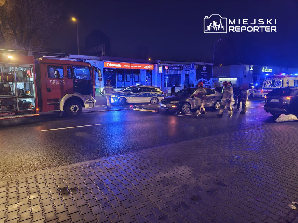 W nocy, strażacy w mundurach podchodzą do samochodu na oświetlonej ulicy, obok policyjny radiowóz.