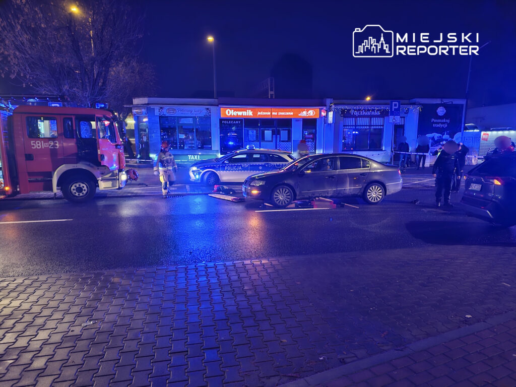 Na ulicy w nocy, strażak w mundurze z urządzeniem do pomiaru, obok policyjny radiowóz i uszkodzony samochód.