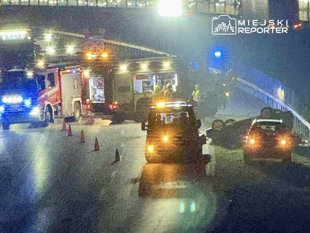 Na jezdni leży przewrócone auto, a służby ratunkowe pracują przy wypadku, oświetlając miejsce akcją.
