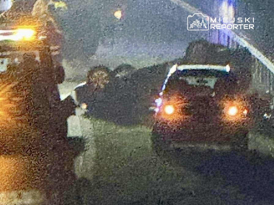 W nocy, na drodze, samochód z włączonymi światłami awaryjnymi stoi obok uszkodzonego pojazdu, a w tle widać służby ratunkowe.