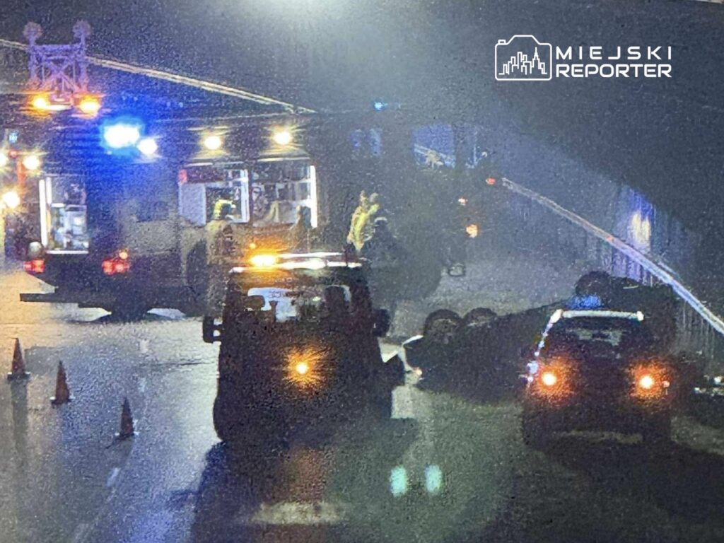 Na drodze widoczna jest przewrócona ciężarówka, a obok pracują strażacy przy wozie strażackim. W tle widać policyjne samochody.