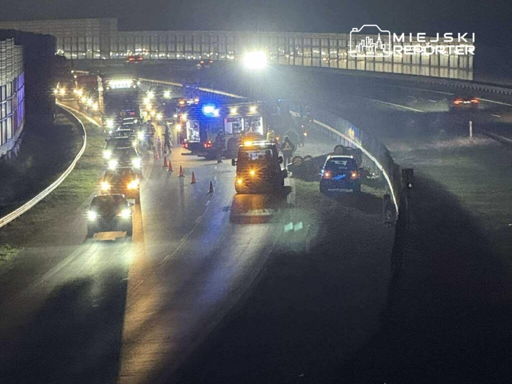 Na drodze nocą trwa akcja ratunkowa po wypadku; widoczne pojazdy służb ratunkowych oraz zablokowane samochody.