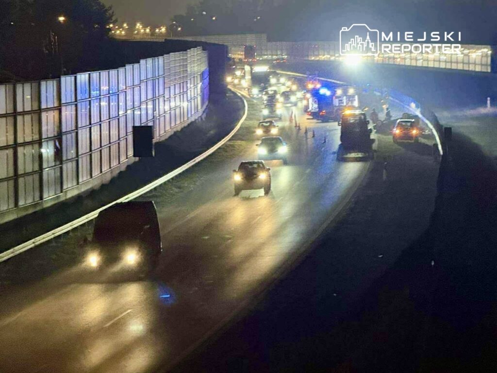 Na drodze nocą widoczne są pojazdy z włączonymi światłami, a w tle pracują służby ratunkowe.