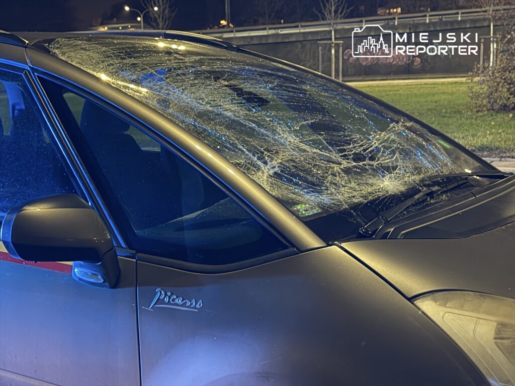 Samochód z pękniętą szybą przednią stoi na parkingu w nocy, oświetlony niebieskim światłem.