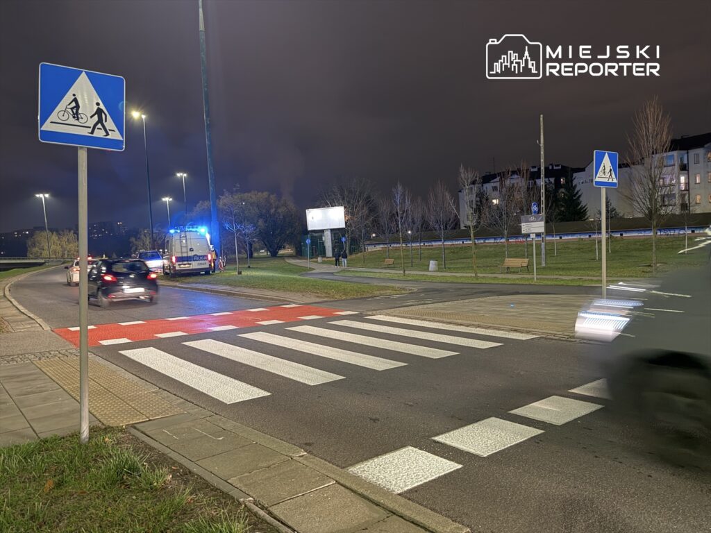 Policyjny radiowóz z włączonymi sygnałami świetlnymi zatrzymany przy przejściu dla pieszych na rondzie w nocy.