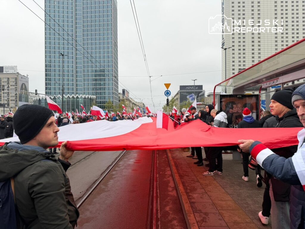 Tłum ludzi trzymający dużą flagę, z budynkami w tle, podczas manifestacji na ulicy.