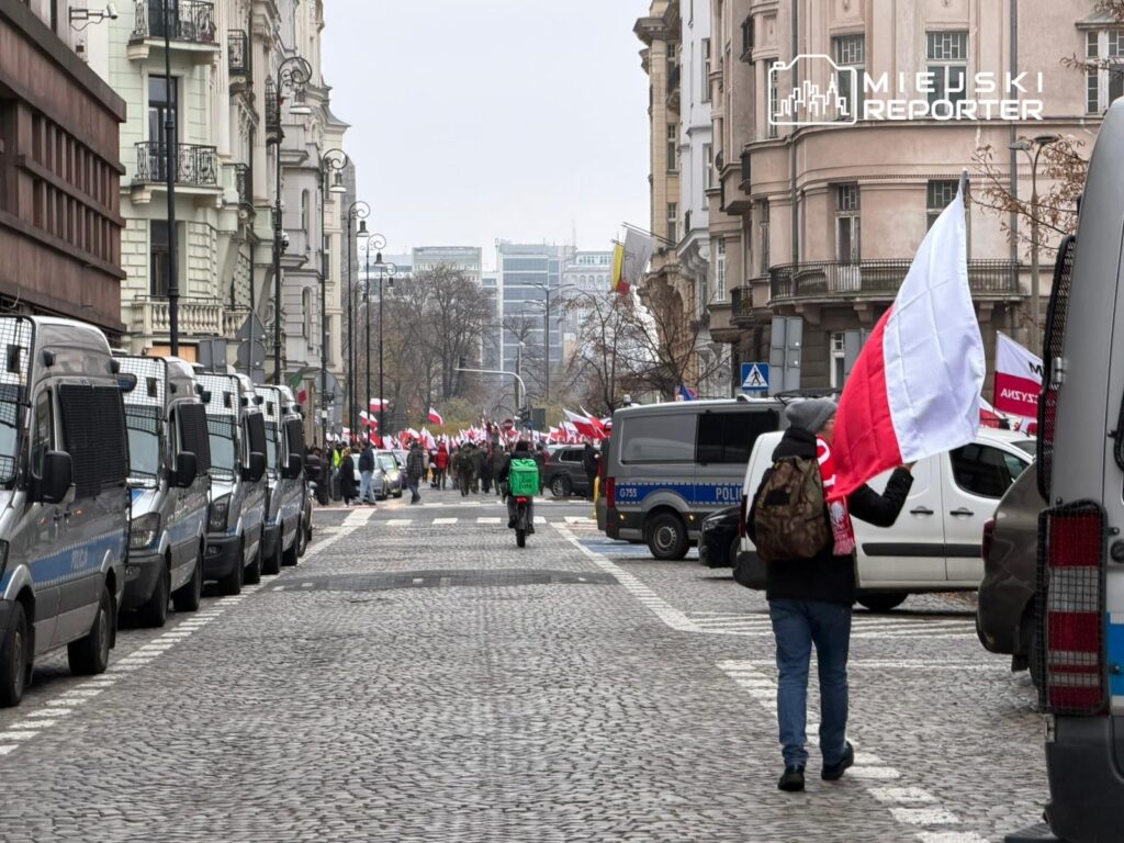 Grupa protestujących z flagami Polski maszeruje ulicą, obok policyjnych furgonetek.