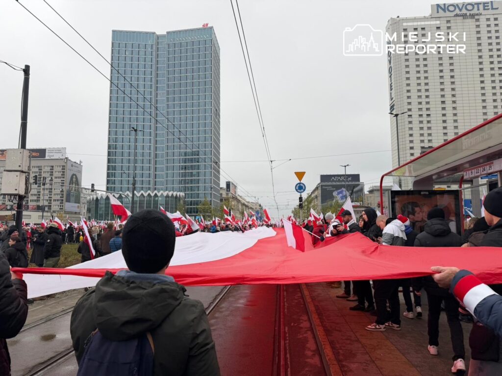 Tłum ludzi trzymających dużą flagę, z nowoczesnymi budynkami w tle, podczas manifestacji w mieście.