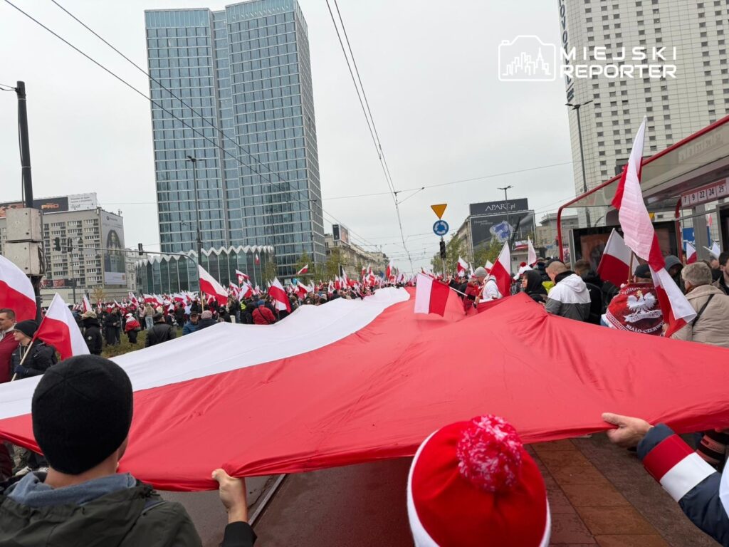 Tłum ludzi trzymający dużą flagę w barwach narodowych na ulicy, w tle nowoczesne budynki i tramwaje.