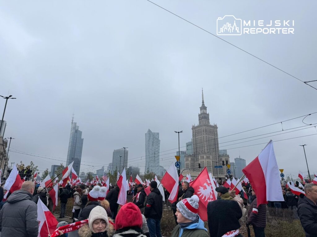 Tłum ludzi z flagami w polskich barwach narodowych gromadzi się na placu, w tle widoczny nowoczesny wieżowiec.