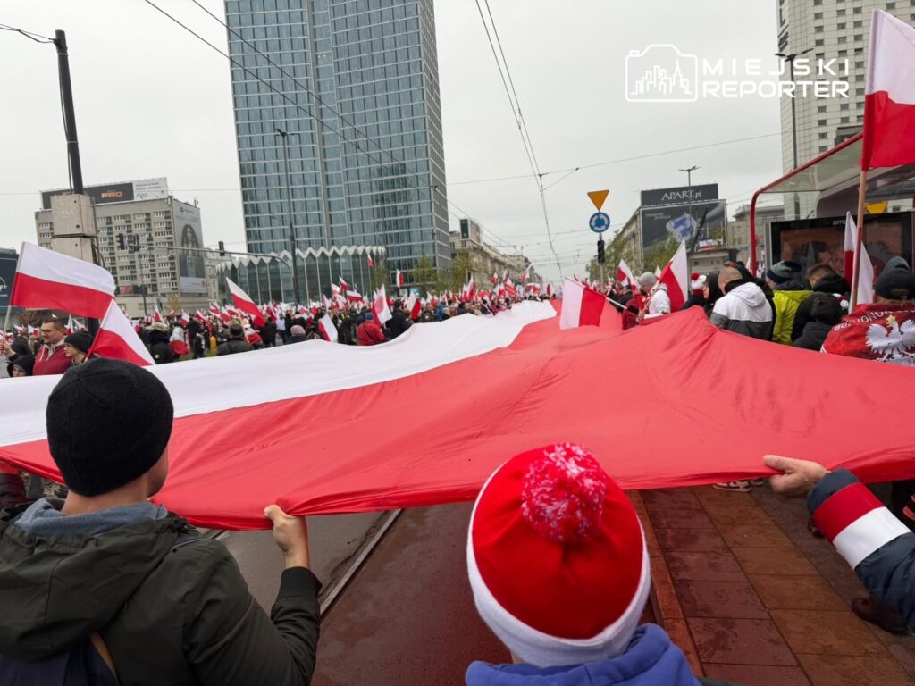 Tłum ludzi trzymający dużą flagę Polski na ulicy, w tle nowoczesne budynki i flagi w rękach uczestników.