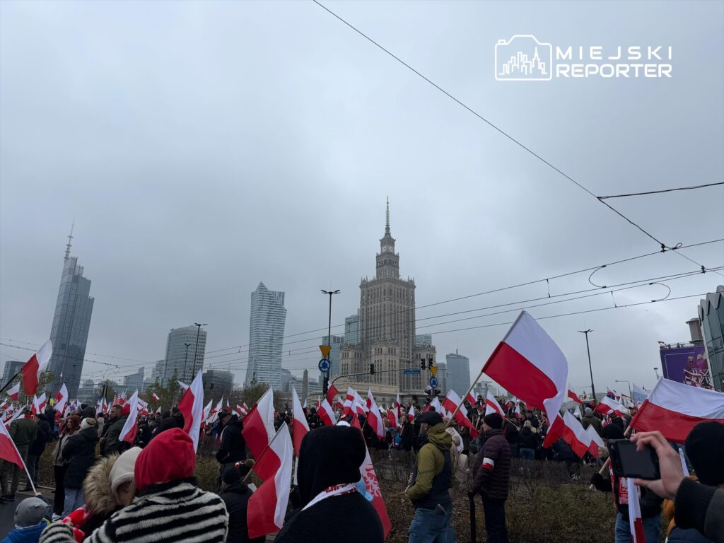 Tłum ludzi z flagami w barwach narodowych zbiera się w miejskiej przestrzeni, z widokiem na wysokie budynki i chmurę.