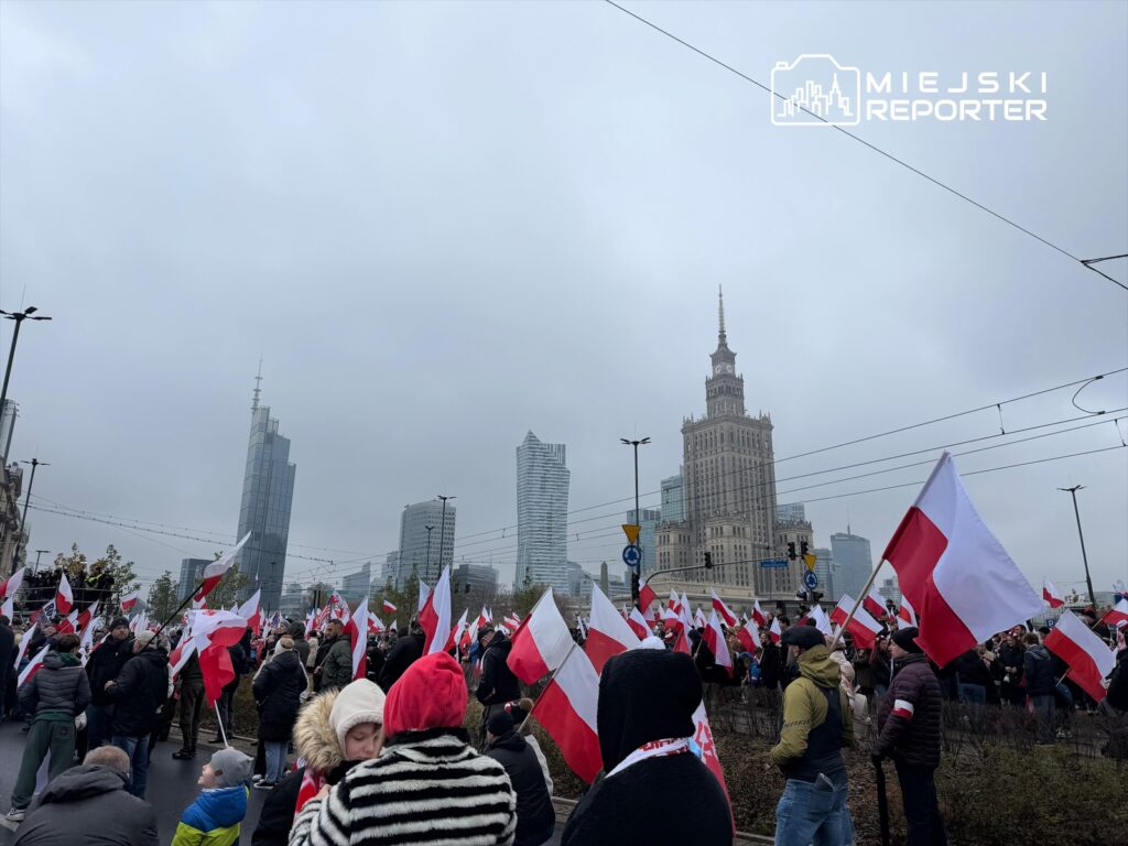 Tłum ludzi trzymających flagi w barwach narodowych, z nowoczesnymi budynkami w tle i szarym niebem.