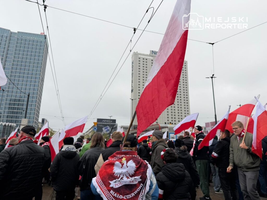 Tłum ludzi z flagami Polski w tle nowoczesnych budynków, uczestniczących w wydarzeniu na ulicy.