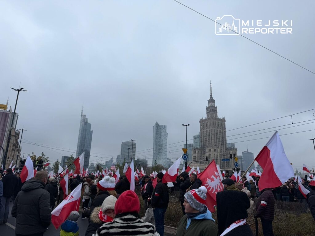 Tłum ludzi z flagami w barwach narodowych zgromadzony na ulicy w mieście, w tle nowoczesne wieżowce oraz historyczny budynek.