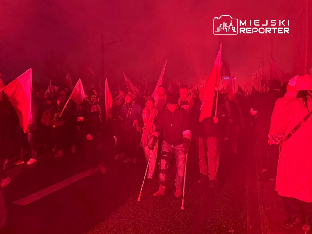 Tłum ludzi niosących flagi w czerwonym świetle, uczestniczący w marszu na ulicy.