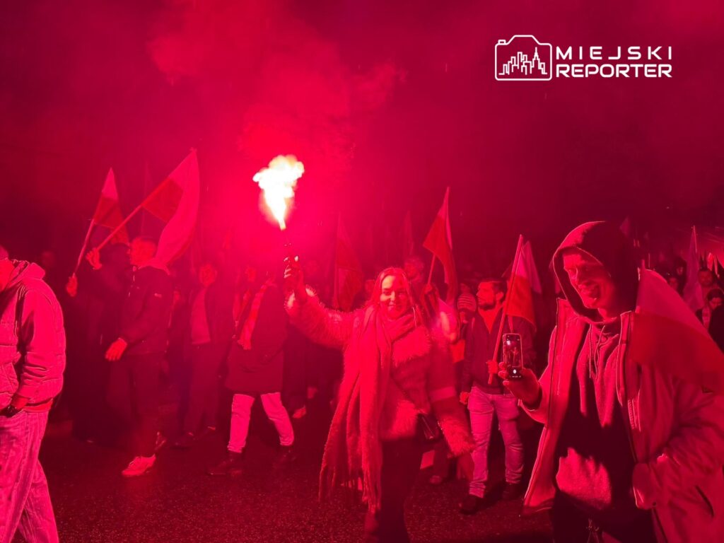 Tłum ludzi z flagami w rękach, przemierzających ulicę w czerwonym świetle rac.