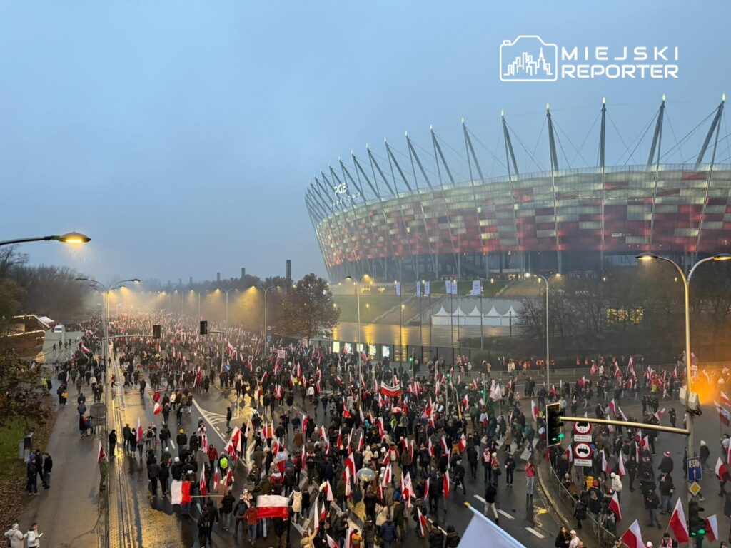 Tłum ludzi z flagami narodowymi przechodzi ulicą obok nowoczesnego stadionu, w tle szare niebo.