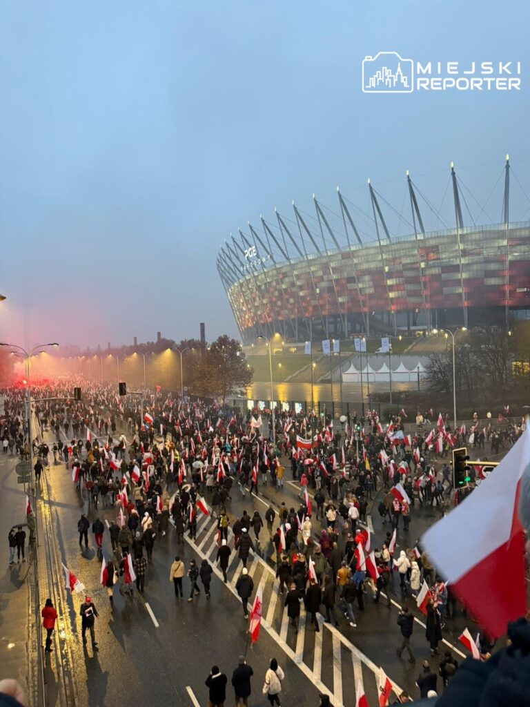Tłum ludzi z flagami narodowymi maszeruje w deszczowej aurze przed nowoczesnym stadionem.