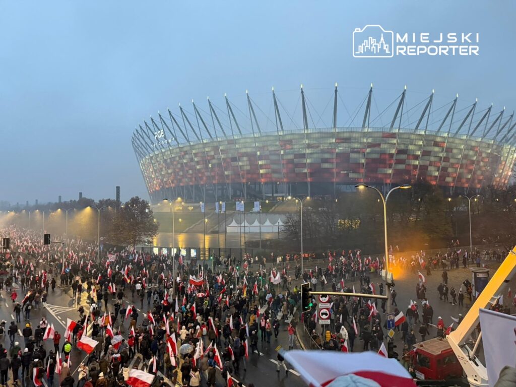 Tłum ludzi z flagami w rękach uczestniczy w marszu przed nowoczesnym stadionem w mgle.
