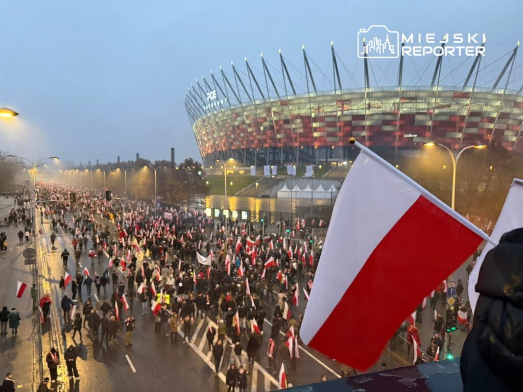 Tłum ludzi niosących flagi w barwach narodowych na ulicy przed nowoczesnym stadionem.
