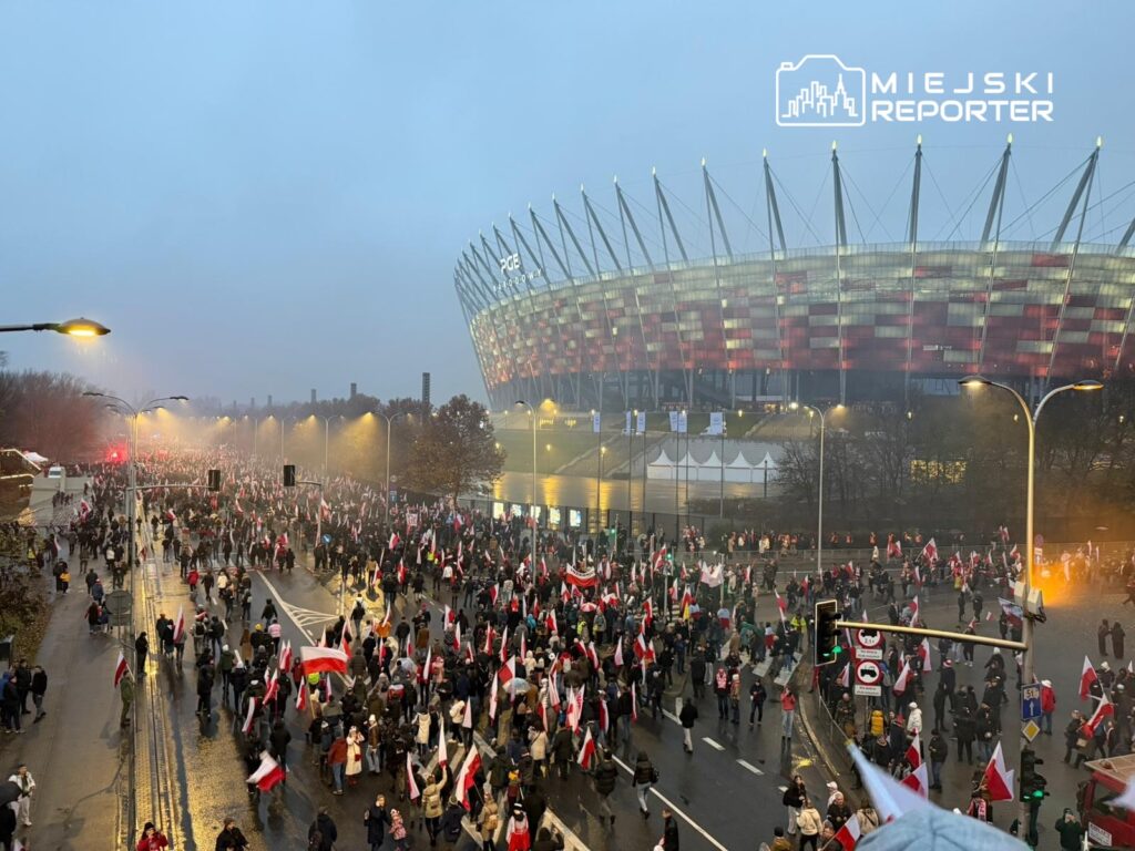 Tłum ludzi z flagami maszeruje w kierunku stadionu, otoczeni mgłą i deszczem.