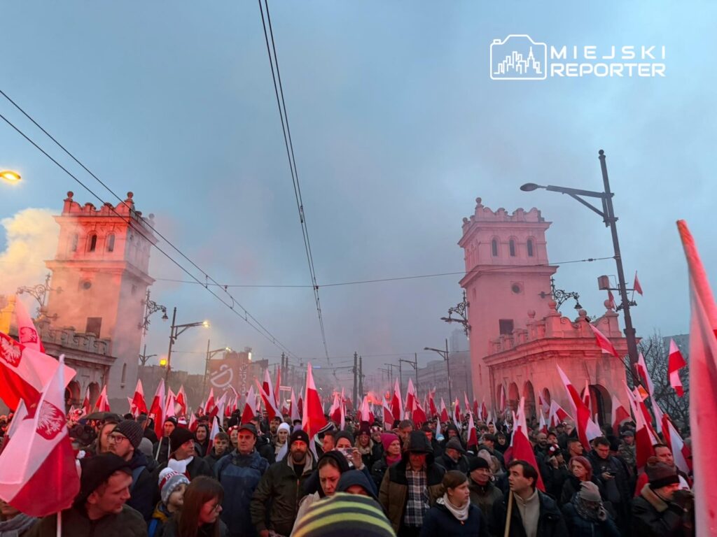 Tłum ludzi z flagami narodowymi wznoszący okrzyki przed historycznymi budynkami w zamglonym otoczeniu.