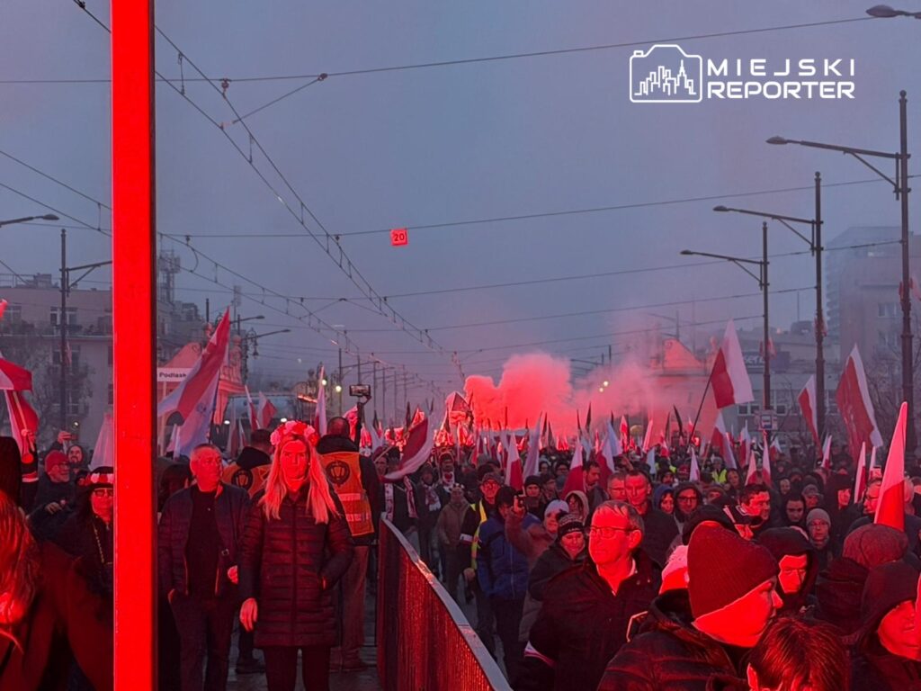 Tłum ludzi z flagami narodowymi maszerujący w kierunku oświetlonym czerwonym światłem, w tle dym.