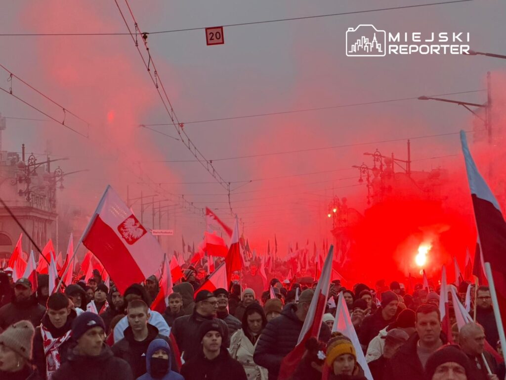 Tłum ludzi z flagami Polski w dymie, uczestniczący w marszu ulicznym w szarej scenerii.