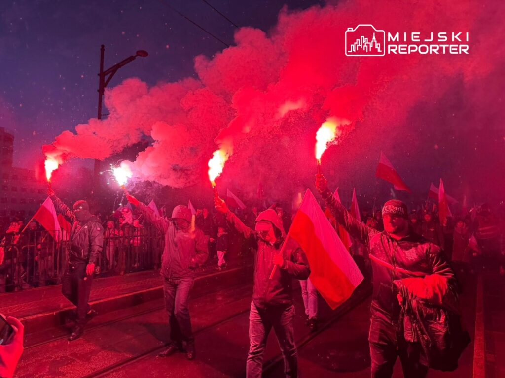Grupa ludzi trzymających flagi, maszerująca w nocnym świetle, trzymająca race, które emitują czerwony dym.