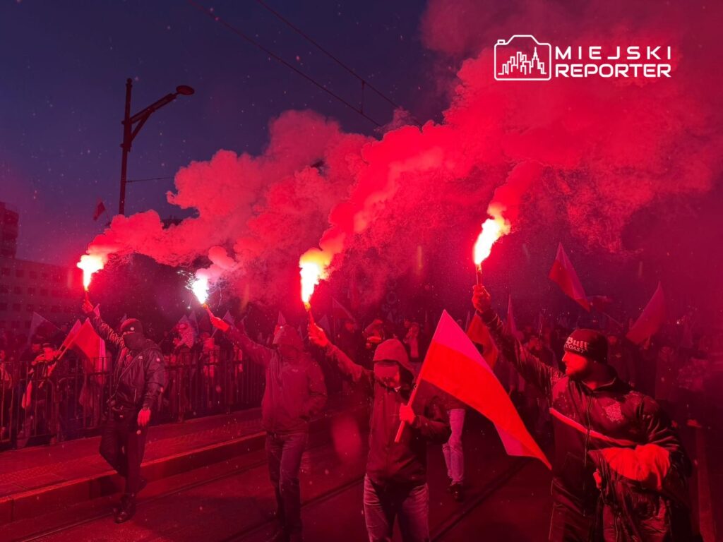 Tłum ludzi trzymających flary i flagi w nocnym świetle, otoczeni chmurami dymu.