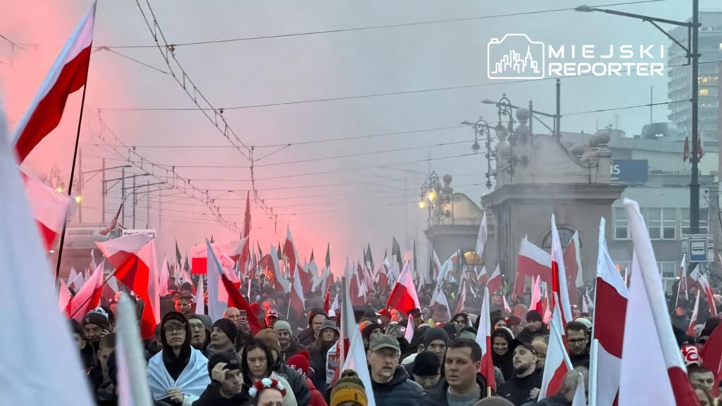 Tłum ludzi z flagami w barwach narodowych maszeruje w dymie na ulicy, otoczeni lampami ulicznymi.