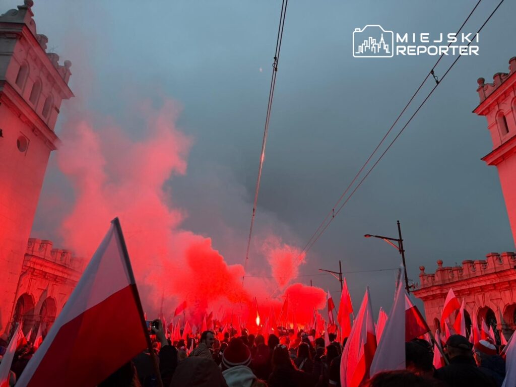Tłum ludzi z flagami w barwach narodowych, otoczonych czerwonym dymem, na ulicy w miejskim otoczeniu.