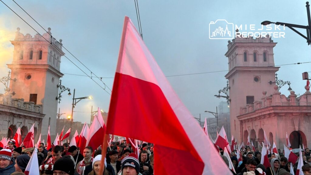 Tłum ludzi z flagami w barwach narodowych przechodzi przed historycznym budynkiem, w tle widoczne dymy.