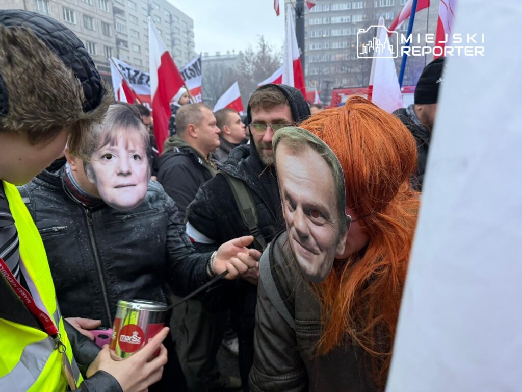Grupa osób w maskach przedstawiających znane twarze uczestniczy w demonstracji, trzymając flagi.