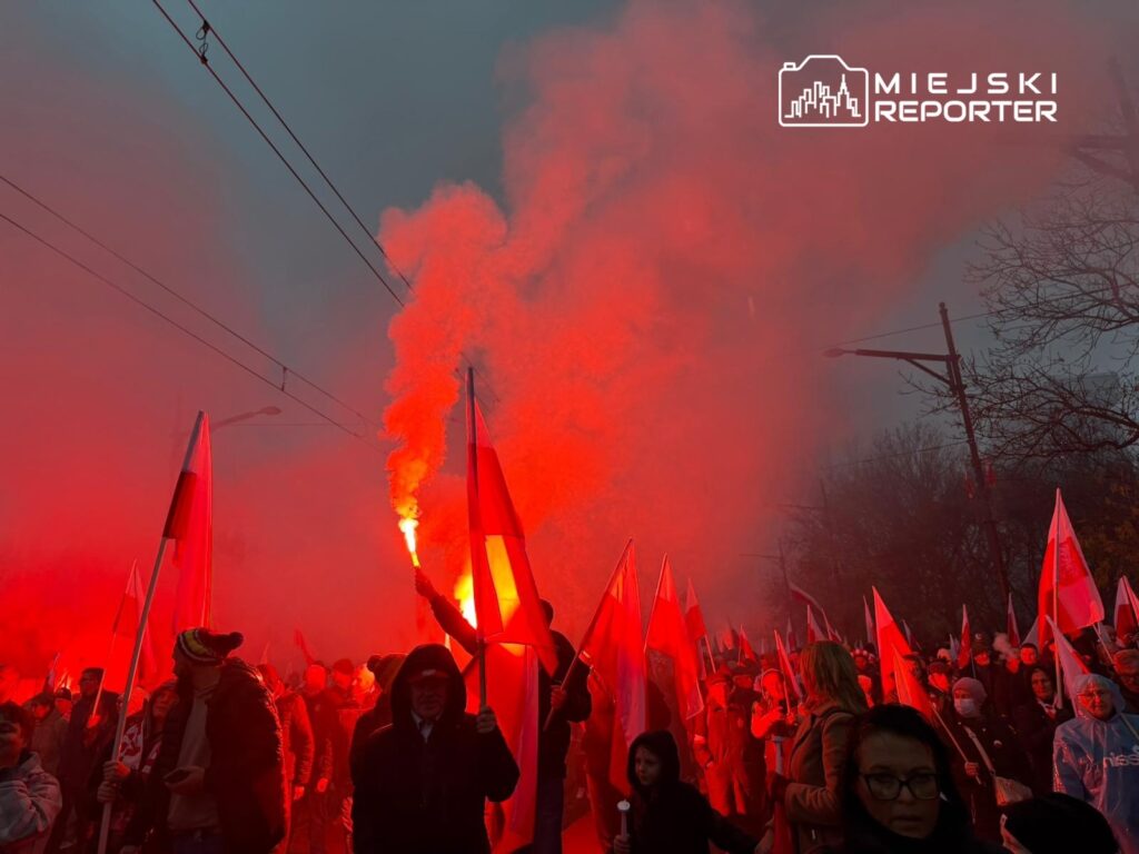 Tłum ludzi z flagami w rękach, unoszący race w czerwonym świetle, na ulicy w mglistym otoczeniu.
