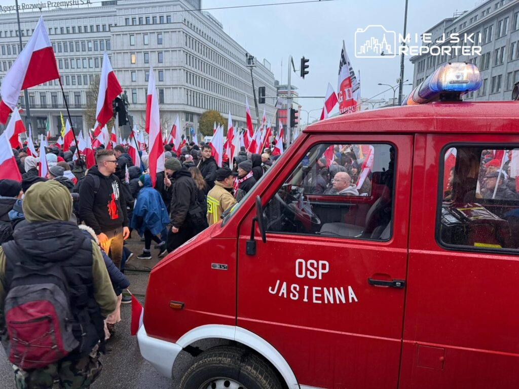 Ludzie trzymający flagi Polski uczestniczą w demonstracji przy czerwonym wozie strażackim OSP Jasienna.