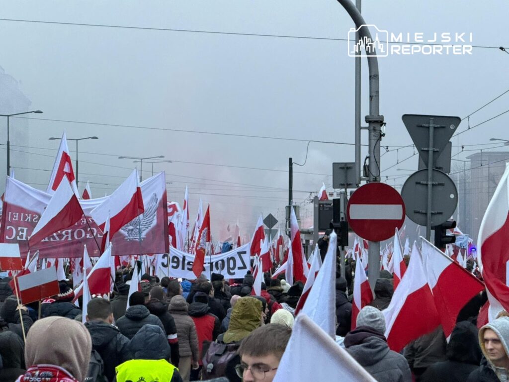 Tłum ludzi z flagami w barwach narodowych idzie ulicą, w tle widoczne znaki drogowe i mglista atmosfera.